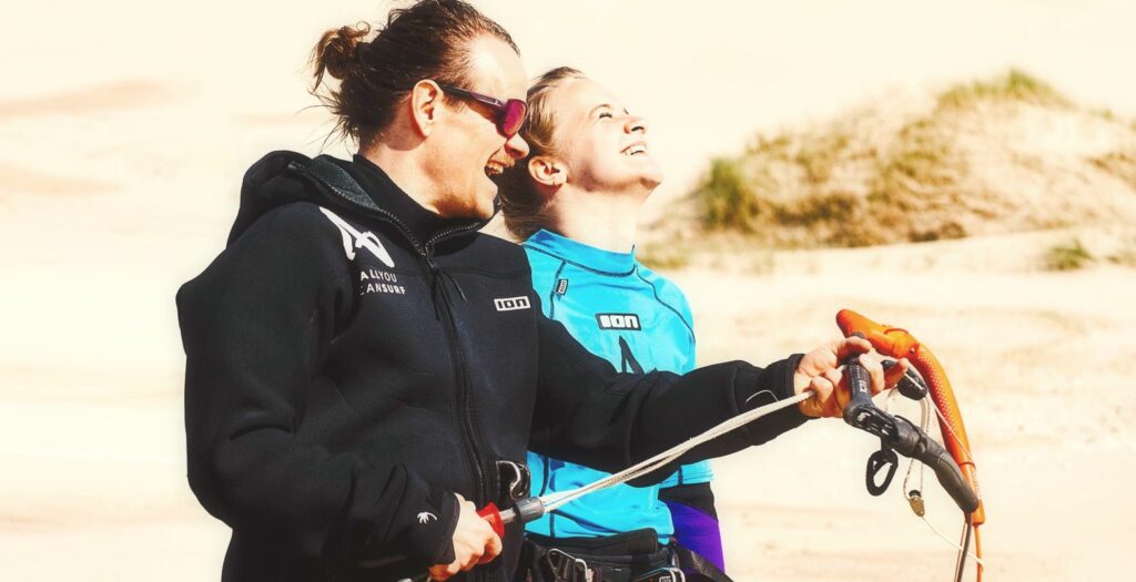 Kitelehrer und lachende Anfaengerin am Strand von Petten, beide mit Blick zum Kite, Hand an der Bar